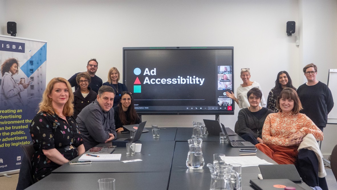A group of 11 alliance members are in a board room smiling for the camera. A big screen behind them shows the Ad Accessibility Alliance logo and three other people in an online meeting.
