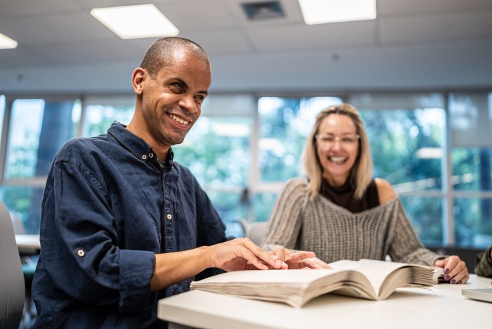 A mid adult man with a visual disability is reading a braille book while a colleague is sat next to him.