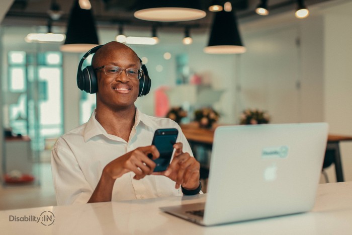 A man sits in front of a laptop with headphones on smiling while also looking at his phone.