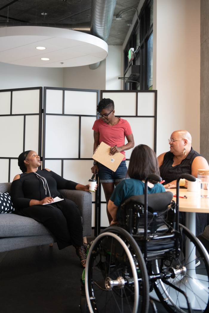 Four disabled people of colour gather around a table during a meeting. A Black woman sitting on a couch gestures and speaks while the three others (a South Asian person sitting in a wheelchair, a Black non-binary person sitting in a chair, and a Black non