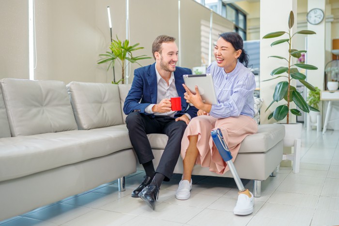 An Asian female worker with a prosthetic leg is having a conversation with her colleague and using a tablet during a coffee break.
