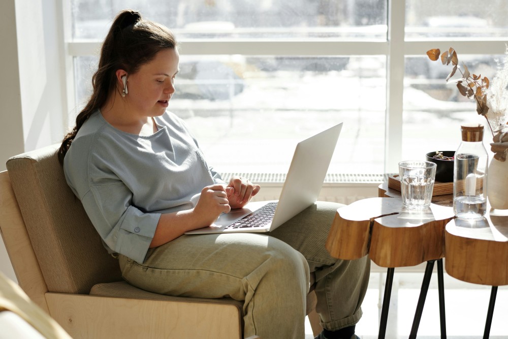 A young woman with Down syndrome is sitting on a chair using a laptop.
