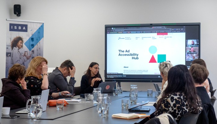 A group of eight alliance members are in a board room in discussion. A big screen behind them shows  three other people in an online meeting.