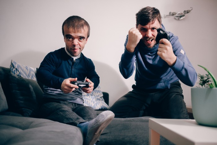 A man with achondroplasia and a friend are playing video games together in a living room.