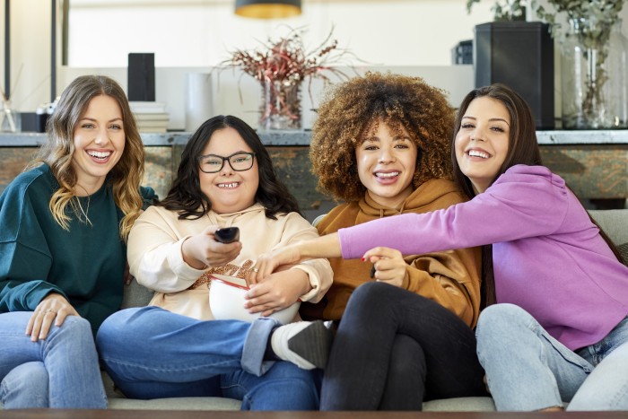 A smiling disabled woman is holding a remote while sitting with female friends on a sofa while holding a bowl of snacks.